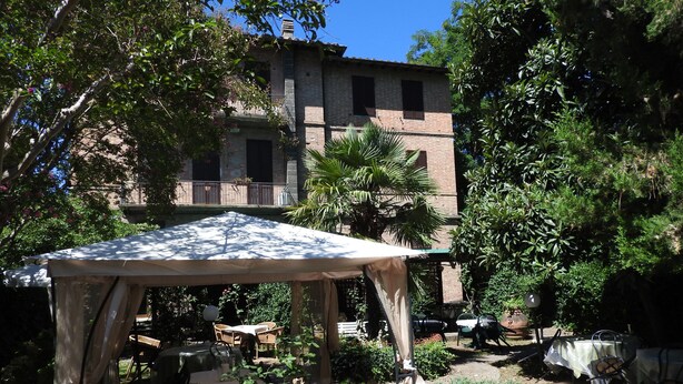 Un espace extérieur abrité, aménagé avec des tables et des chaises, entouré d’une végétation luxuriante et avec un bâtiment en arrière-plan.