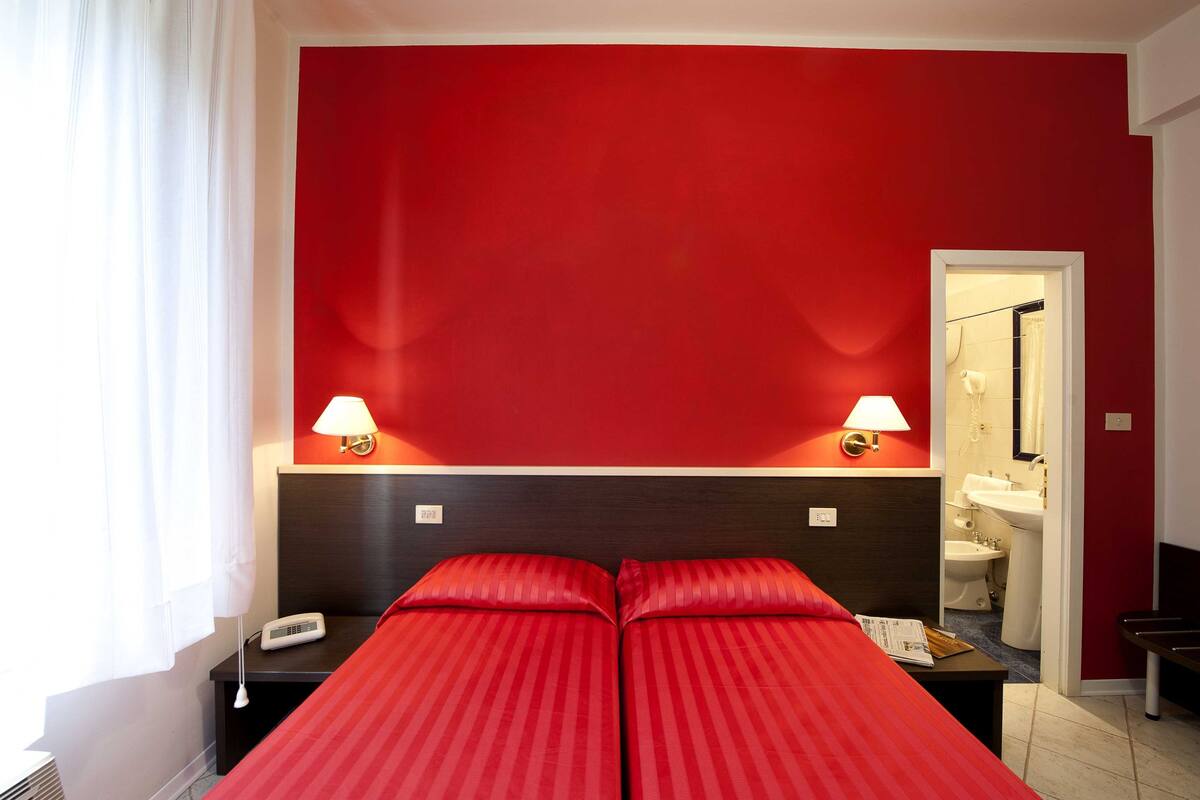 Ein Zimmer mit roter Wand, einem Bett mit roter Bettwäsche, zwei Nachttischlampen, einem Nachttisch und Blick auf ein Badezimmer mit Waschbecken und Toilette.