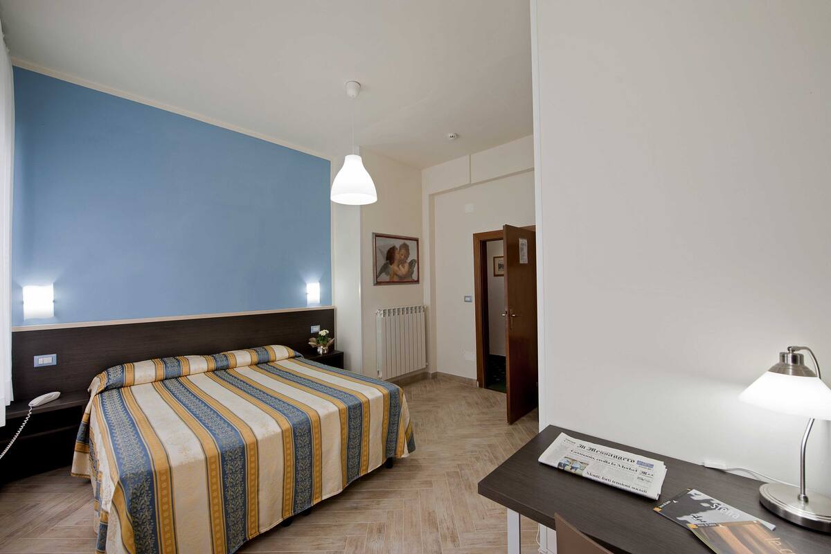 Ein Hotelzimmer mit Bett, Schreibtisch mit Lampe, Telefon und einem gerahmten Bild an der Wand.