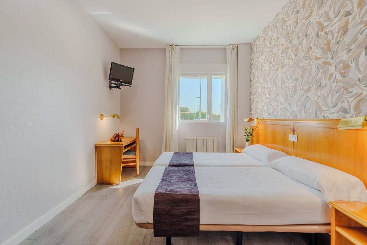 Une chambre d’hôtel avec deux lits, une table de chevet en bois, un bureau, un téléviseur à écran plat fixé au mur et une fenêtre donnant sur l’extérieur.
