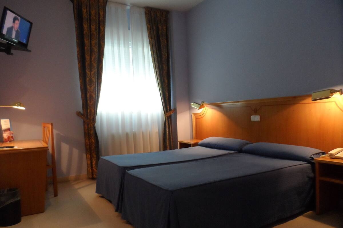 Une chambre d’hôtel avec un lit, un bureau, une chaise, une télévision et une fenêtre avec des rideaux.