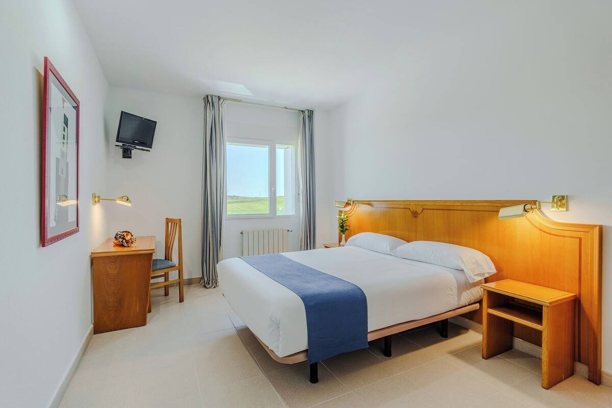 Une chambre d’hôtel comprenant un lit, une table de chevet, une chaise, une télévision, une fenêtre et des rideaux.