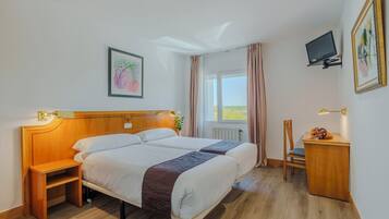 Habitación de hotel con cabecera de madera, dos camas, una mesita pequeña de madera, una silla, un televisor y una ventana con cortinas.