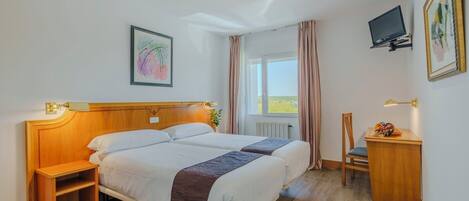 A hotel room with a wooden headboard, two beds, a small wooden table, a chair, a TV, and a window with curtains.