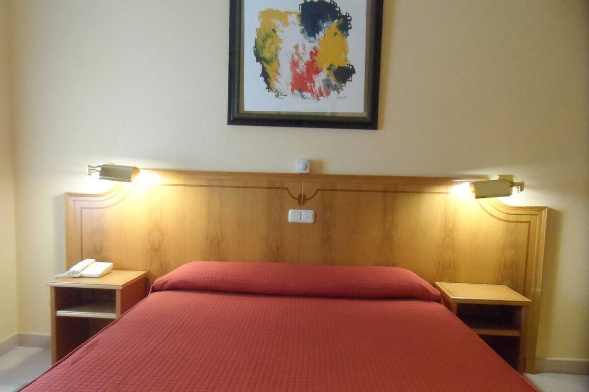 Une chambre d’hôtel avec un grand lit, deux tables de chevet, une petite table d’appoint, un tableau abstrait encadré et une tête de lit en bois.