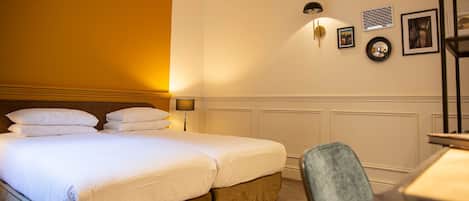 A hotel room with two beds, a desk, and a chair. There is a wall-mounted lamp and framed pictures on the wall.