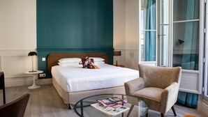 Quarto de hotel com uma cama, uma cadeira, uma mesinha de centro com uma revista, um abajur e uma janela com cortinas.