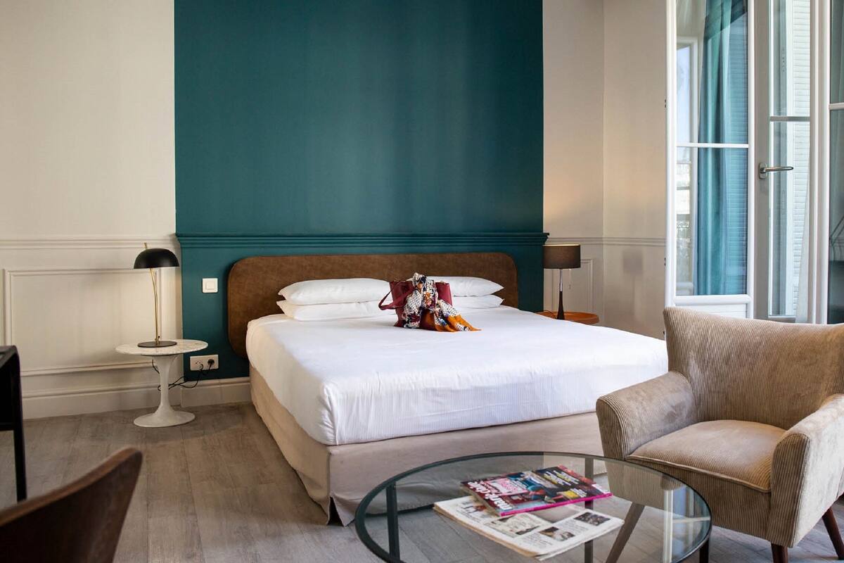 Une chambre d’hôtel avec un lit, une chaise, une petite table avec un magazine, une lampe et une fenêtre avec des rideaux.