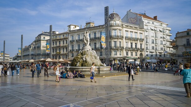 Uma praça pública com uma fonte central, rodeada por edifícios históricos e uma multidão de pessoas.