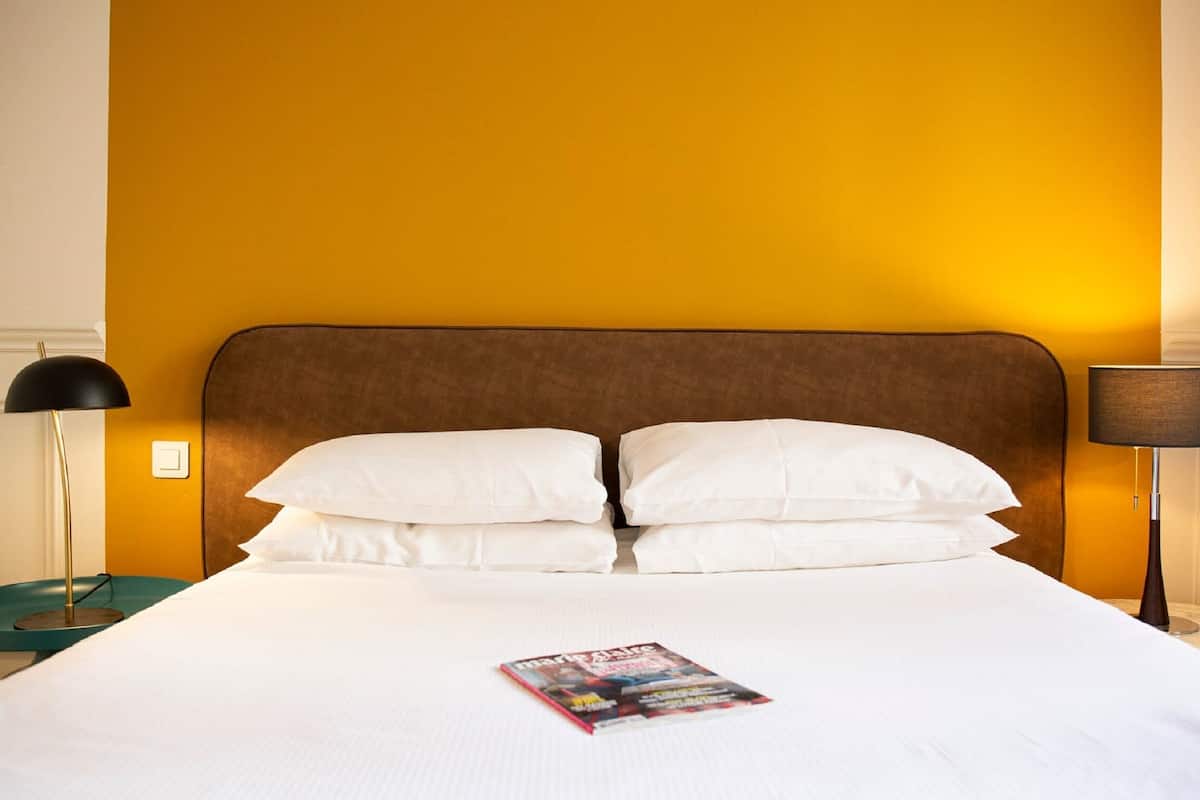 Un lit bien fait, avec du linge de lit blanc et des oreillers, un magazine posé sur le lit, et deux lampes de chevet de chaque côté.