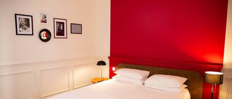 A neatly made bed with white linens, a red accent wall, and a small round table with a lamp.