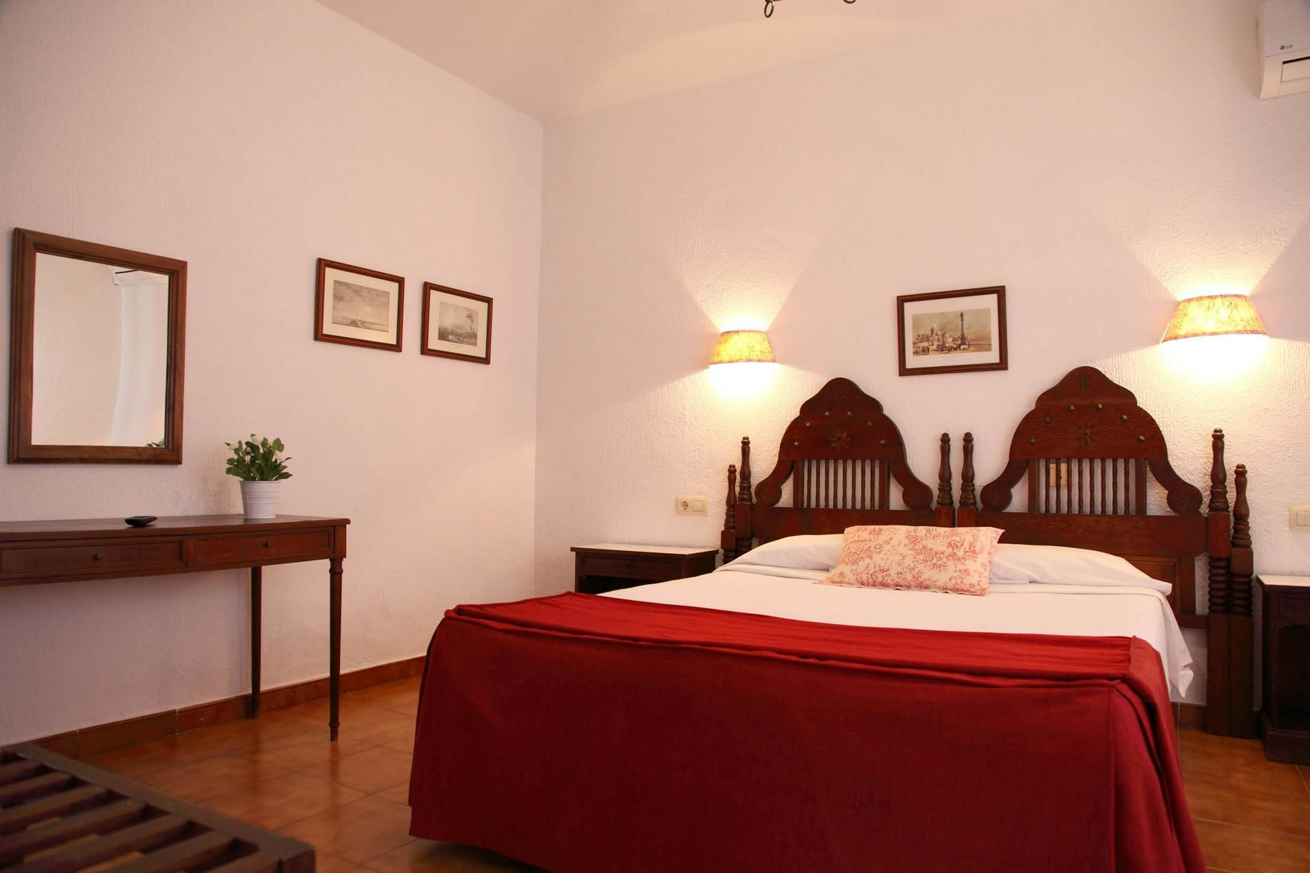 Un dormitorio con una cama grande, una mesita de noche de madera, una mesita auxiliar, un espejo y dos lámparas de pared.