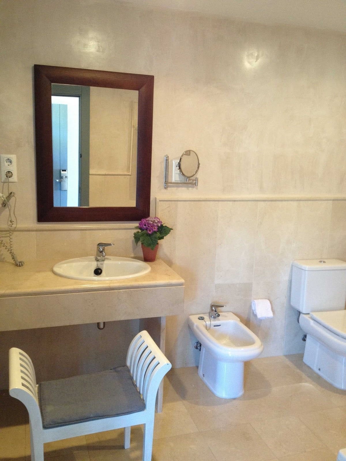 Un baño con lavamanos, espejo, inodoro, bidé y una silla.
