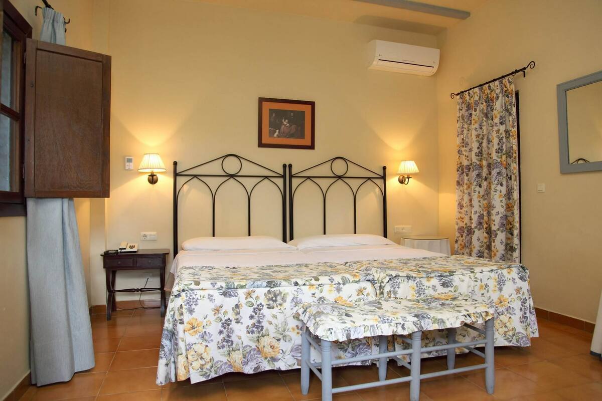 Un dormitorio con una cama, dos mesitas de noche, un armario de madera, un equipo de aire acondicionado, un cuadro enmarcado y una cortina con motivos florales.