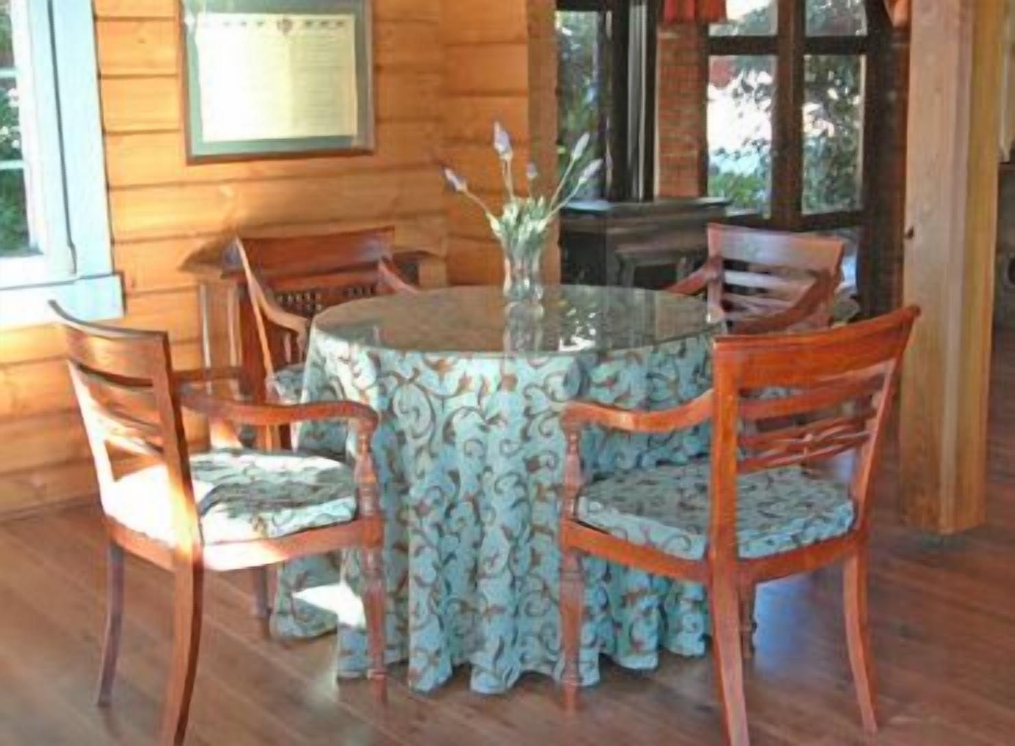 A wooden cabin interior with a round dining table surrounded by four chairs, a vase with flowers, and a window with a view of trees.