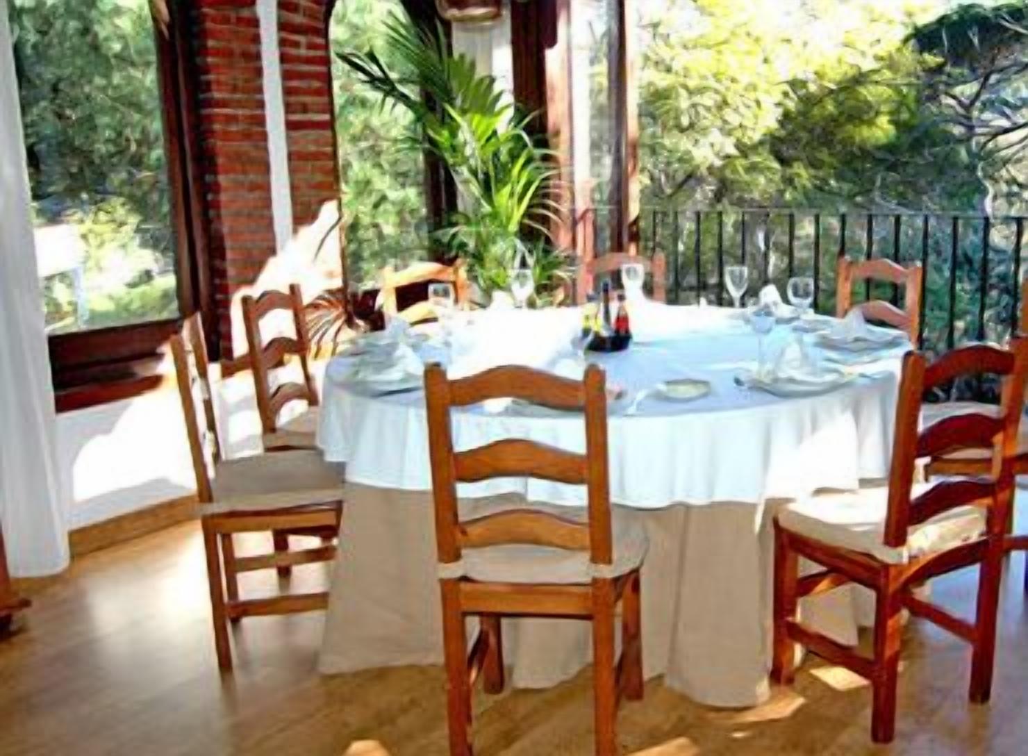 A round table set for dining with wooden chairs, surrounded by a brick wall and large windows.
