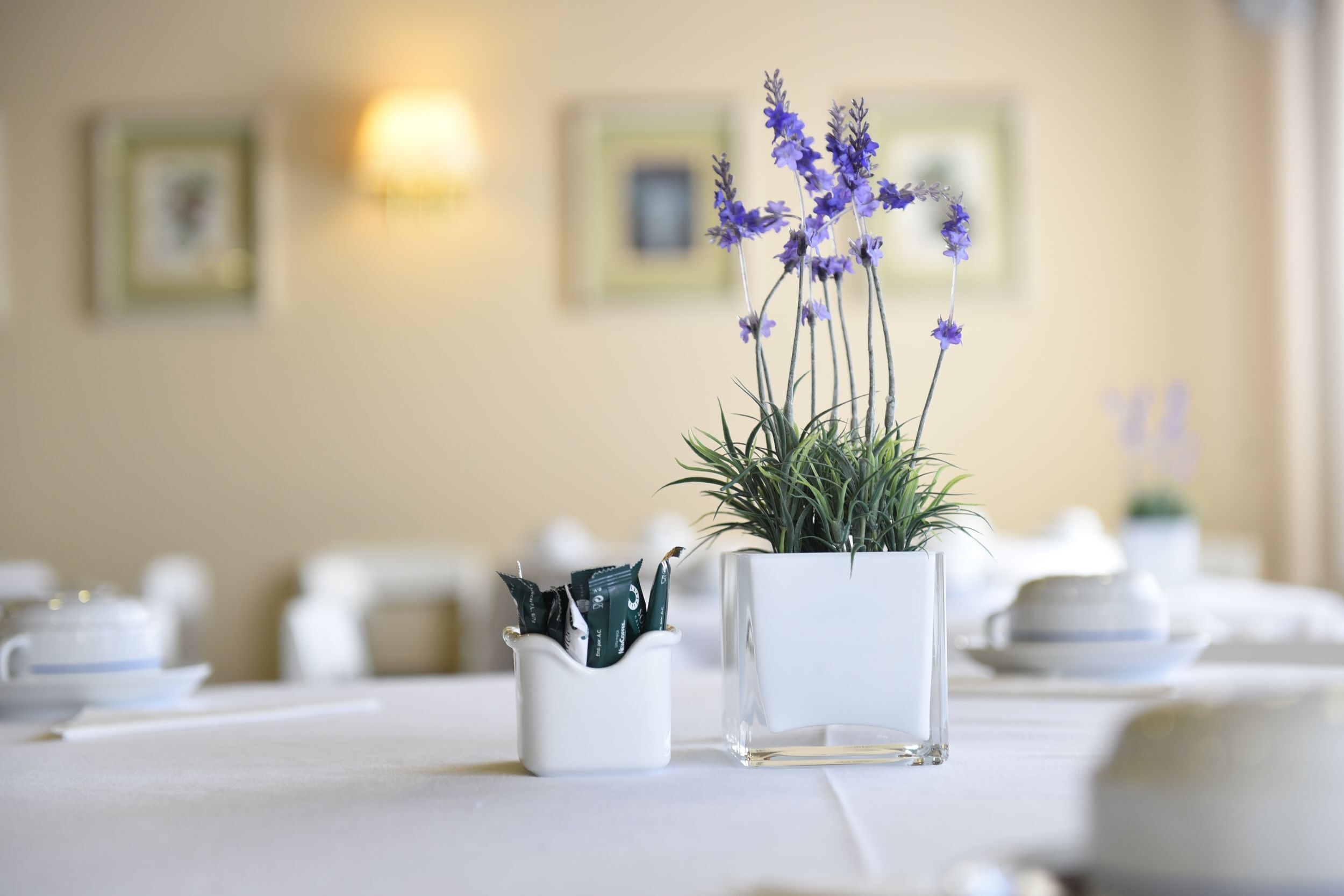Une table à manger ornée d’un vase rempli de fleurs violettes, d’un petit récipient blanc contenant des objets verts et d’un vase en verre transparent avec une plante.