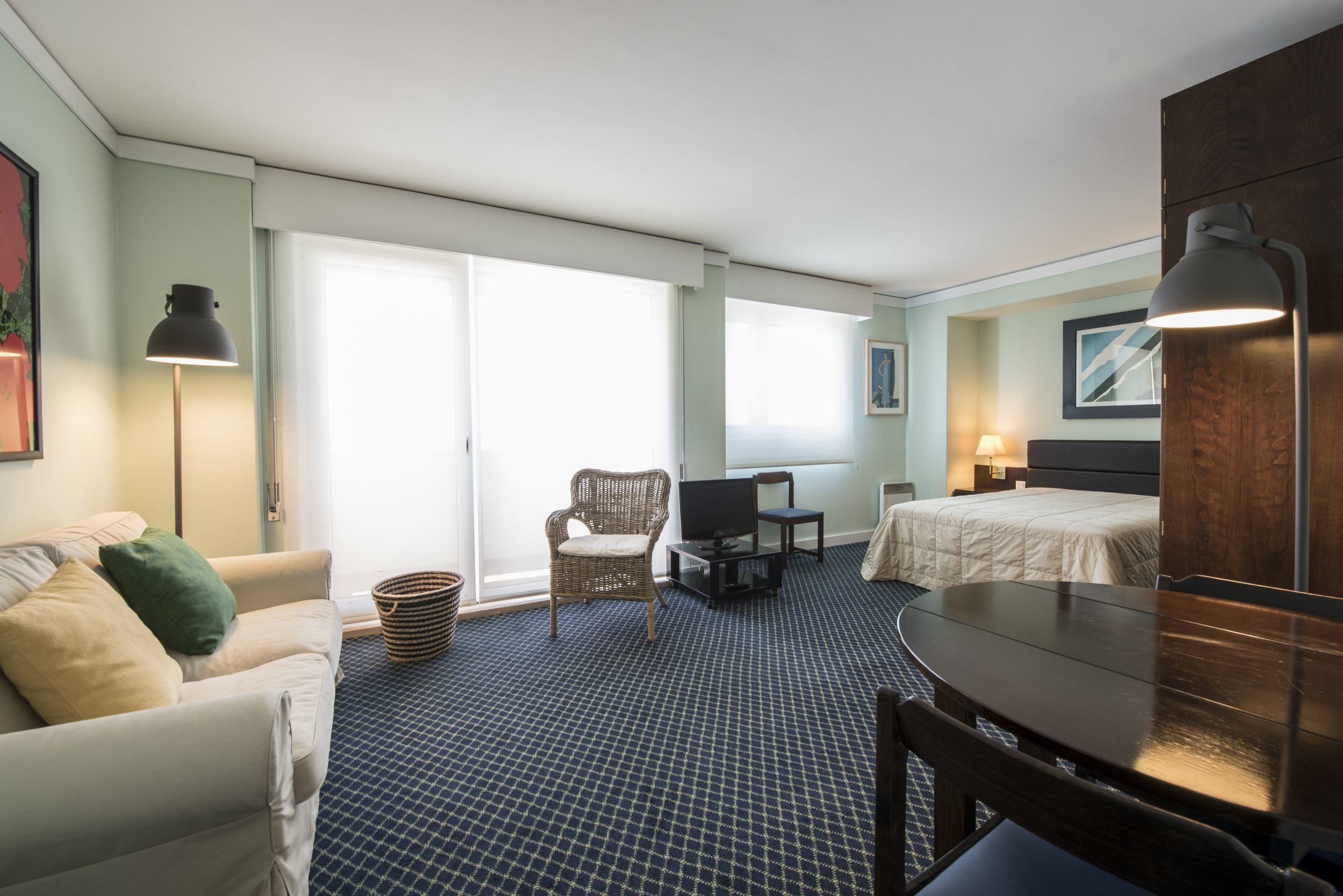 A hotel room with a bed, a sofa, a dining table, a TV, and a lamp.