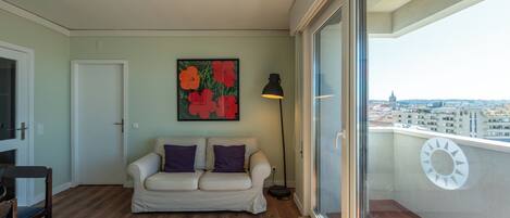 A balcony with a view of a cityscape, a white sofa with purple cushions, and a painting of red flowers on the wall.