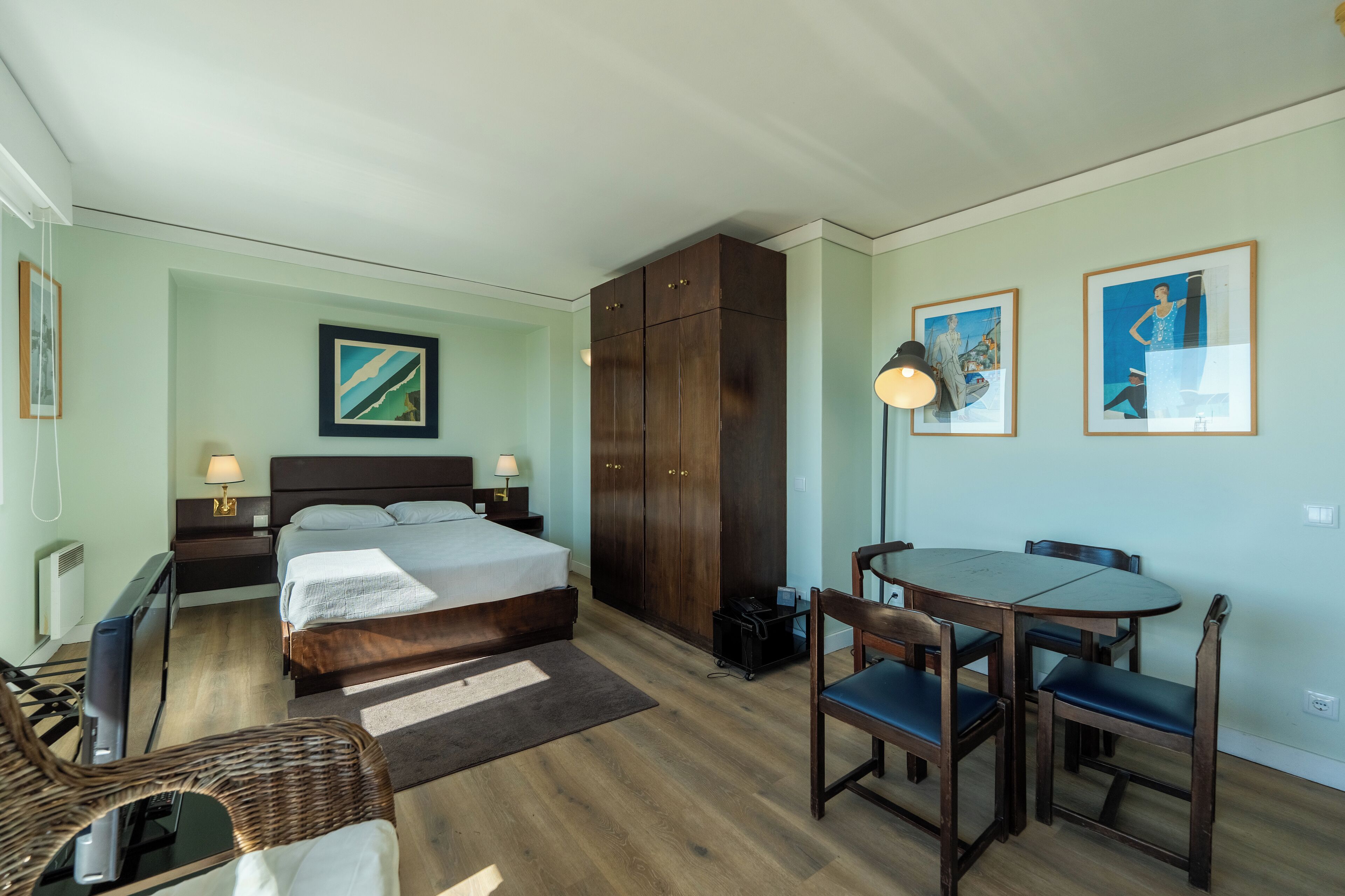 Une chambre d’hôtel comprenant un lit, des tables de chevet, un bureau avec une chaise, une table à manger avec des chaises et des œuvres d’art accrochées aux murs.