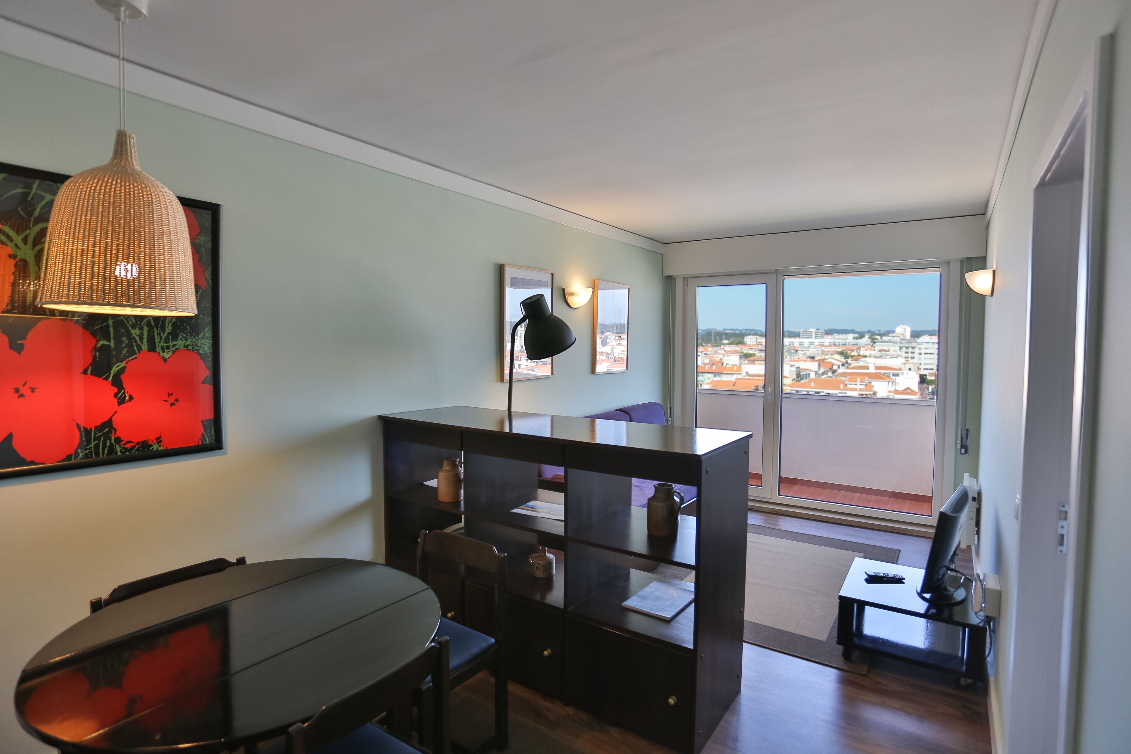 A modern room with a dining table, chairs, a black desk, and a large window offering a city view.
