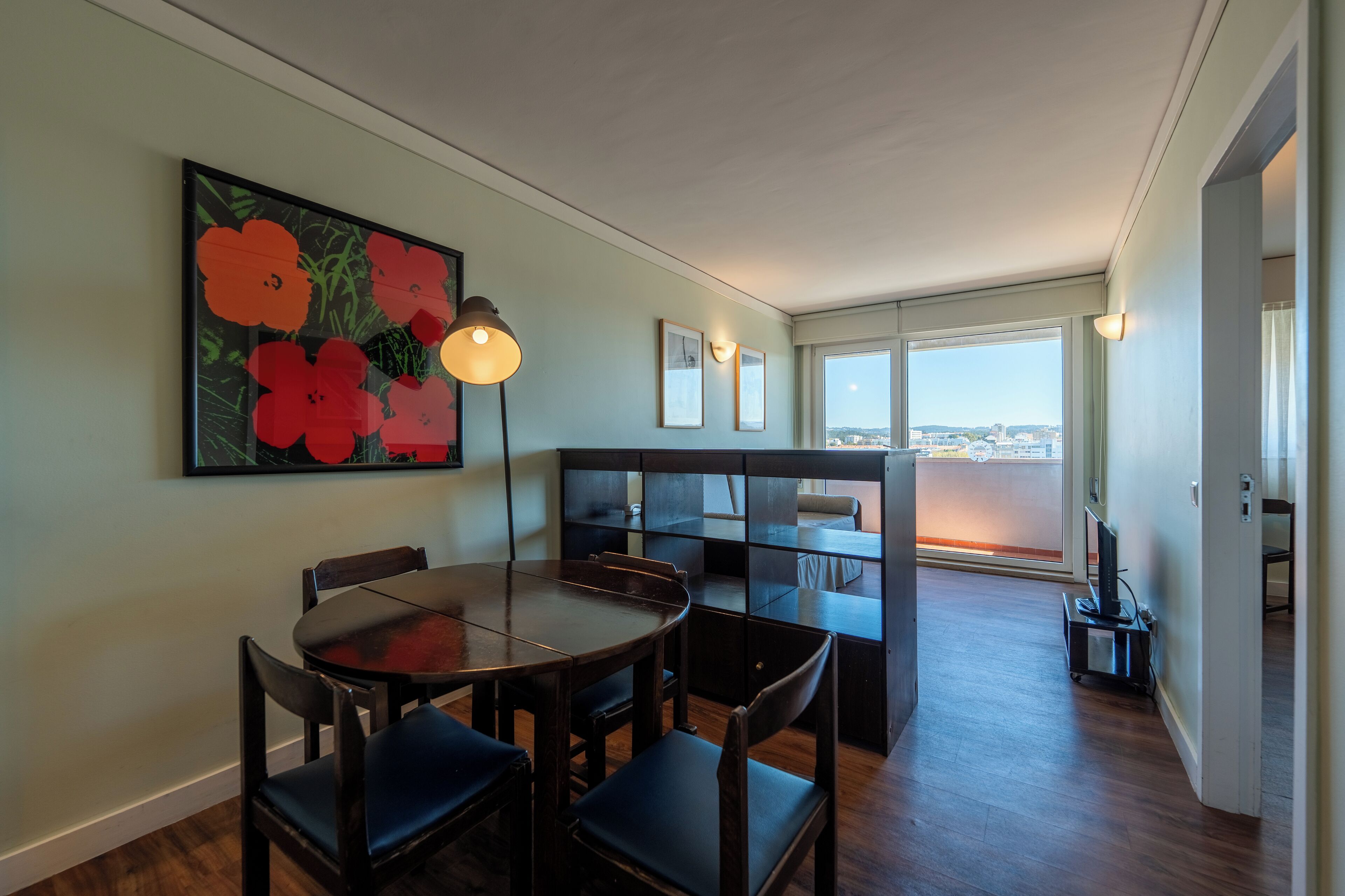 Un coin repas avec une table ronde et des chaises, un tableau de fleurs accroché au mur et une vue sur la ville à travers de grandes fenêtres.