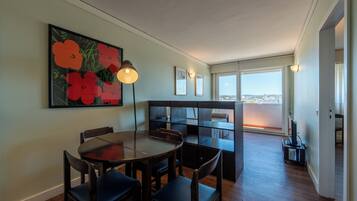Un coin repas avec une table ronde et des chaises, un tableau de fleurs accroché au mur et une vue sur la ville à travers de grandes fenêtres.