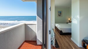 A balcony with a view of the beach and ocean, a bedroom with a bed, a bedside table, and a lamp.