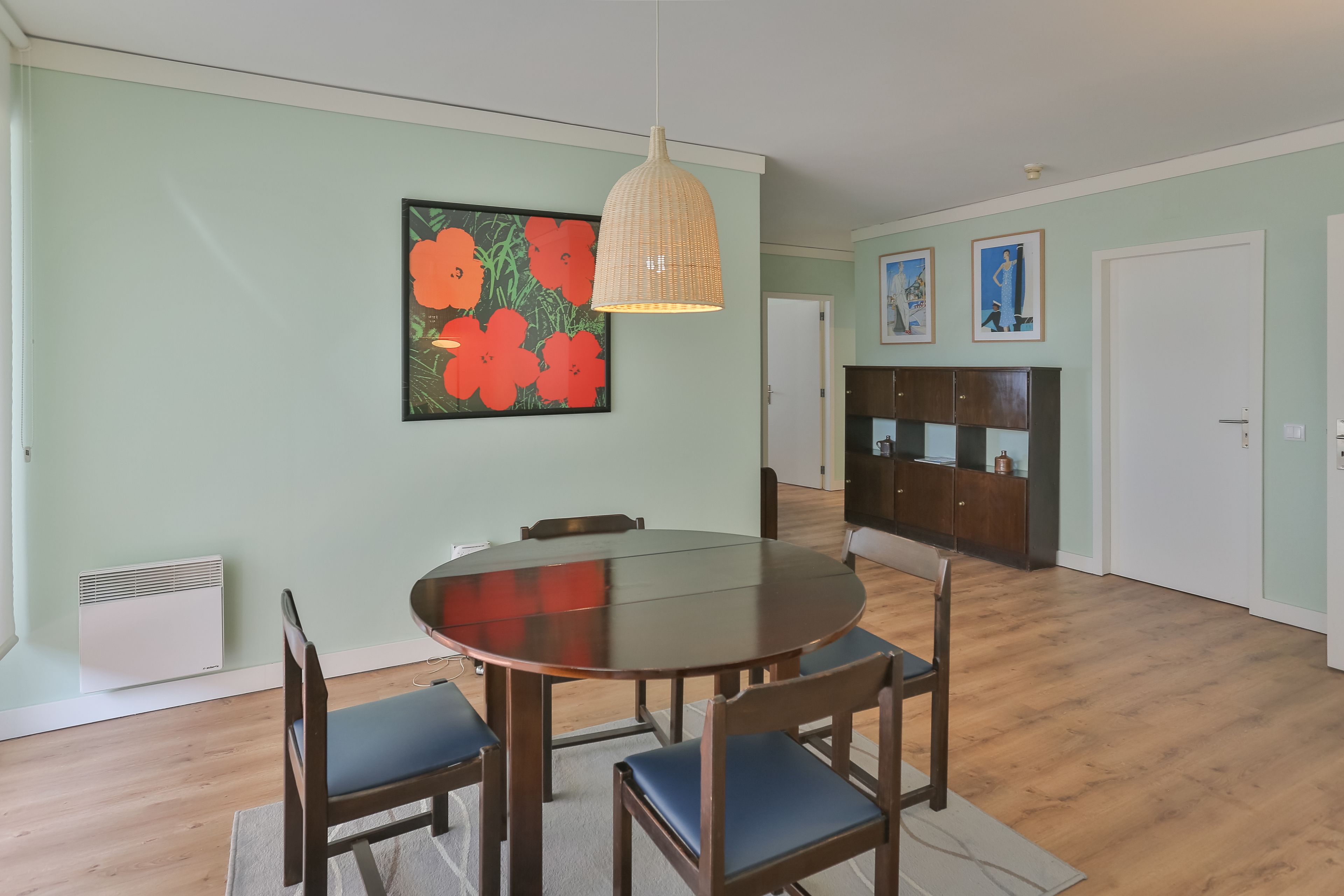Un coin repas comprenant une table ronde, quatre chaises, un luminaire suspendu et un grand tableau représentant des fleurs accroché au mur.