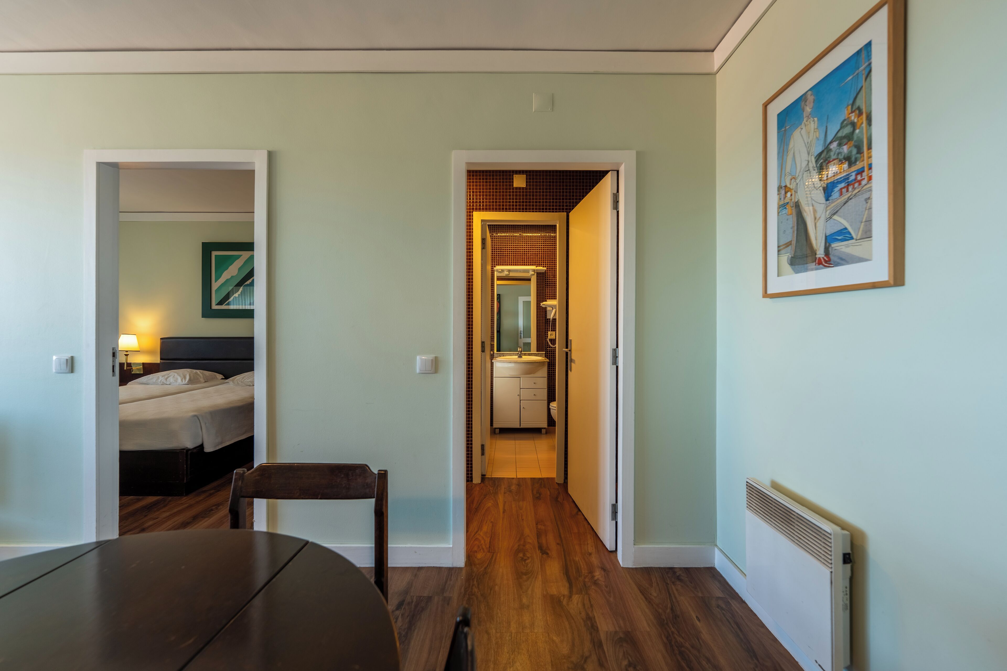 Habitación de hotel con piso de madera, una cama, una silla y una mesa. Se ve un baño a través de una puerta abierta.