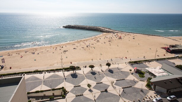 A beach with a paved area in the foreground, a rocky breakwater, and a clear view of the ocean.
