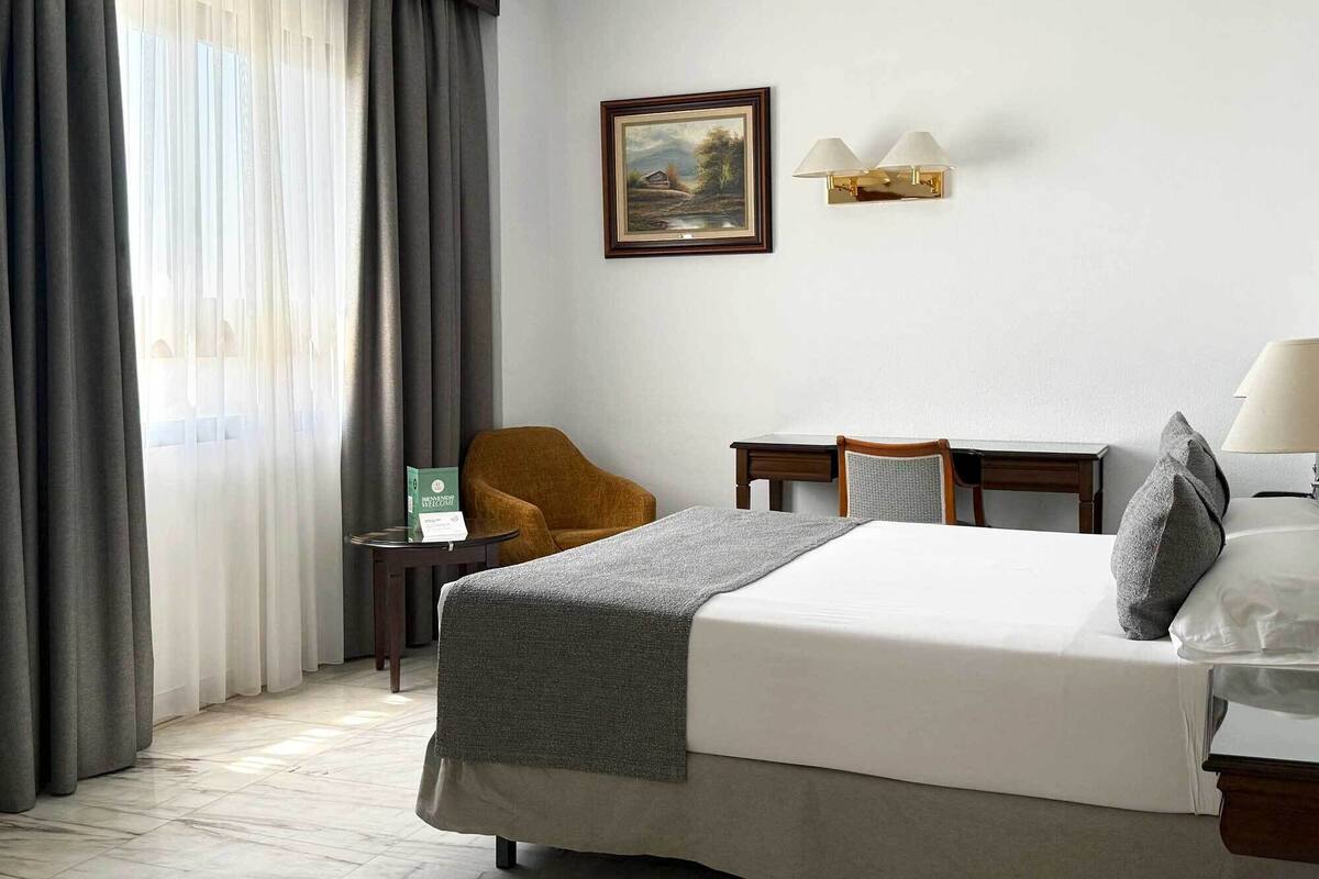 A hotel room with a bed, a desk, a chair, a painting on the wall, and a window with curtains.