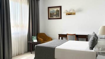 Une chambre d’hôtel avec un lit, un bureau, une chaise, un tableau au mur et une fenêtre avec des rideaux.