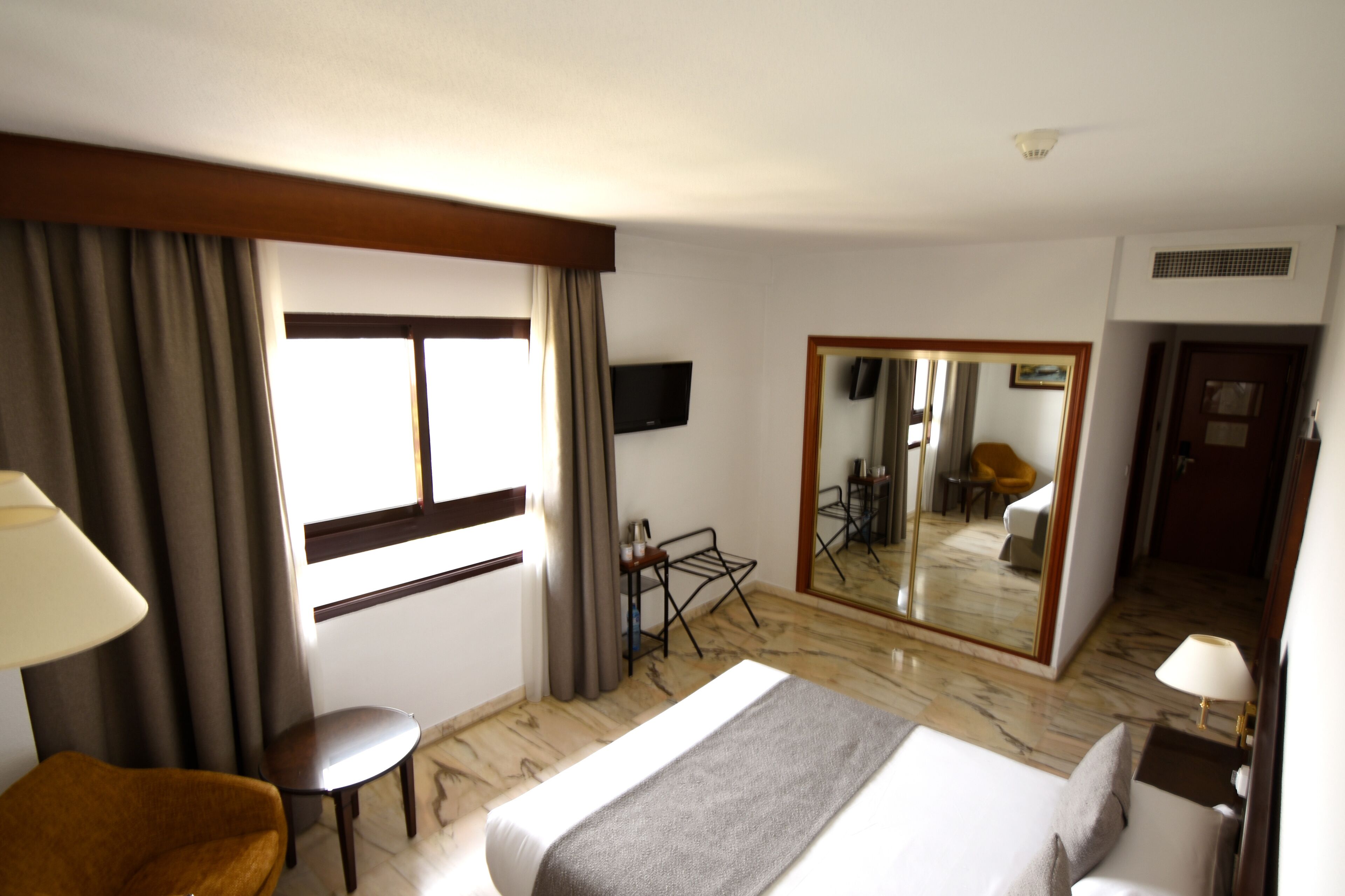 Une chambre d’hôtel équipée d’un lit, d’un bureau, d’une chaise, d’une télévision, d’un miroir et d’une fenêtre avec des rideaux.