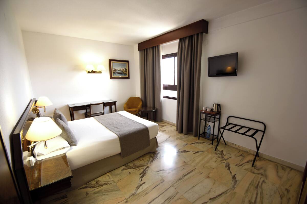 A hotel room with a bed, bedside table, lamp, chair, TV, and a small table with a cup.