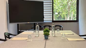 Un salón de conferencias con una gran televisión de pantalla plana, una ventana que ofrece vistas a árboles, y una mesa dispuesta con cristalería y una pequeña planta.