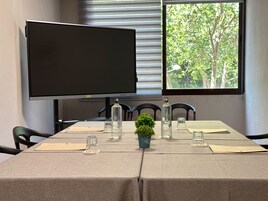 Un salón de conferencias con una gran televisión de pantalla plana, una ventana que ofrece vistas a árboles, y una mesa dispuesta con cristalería y una pequeña planta.