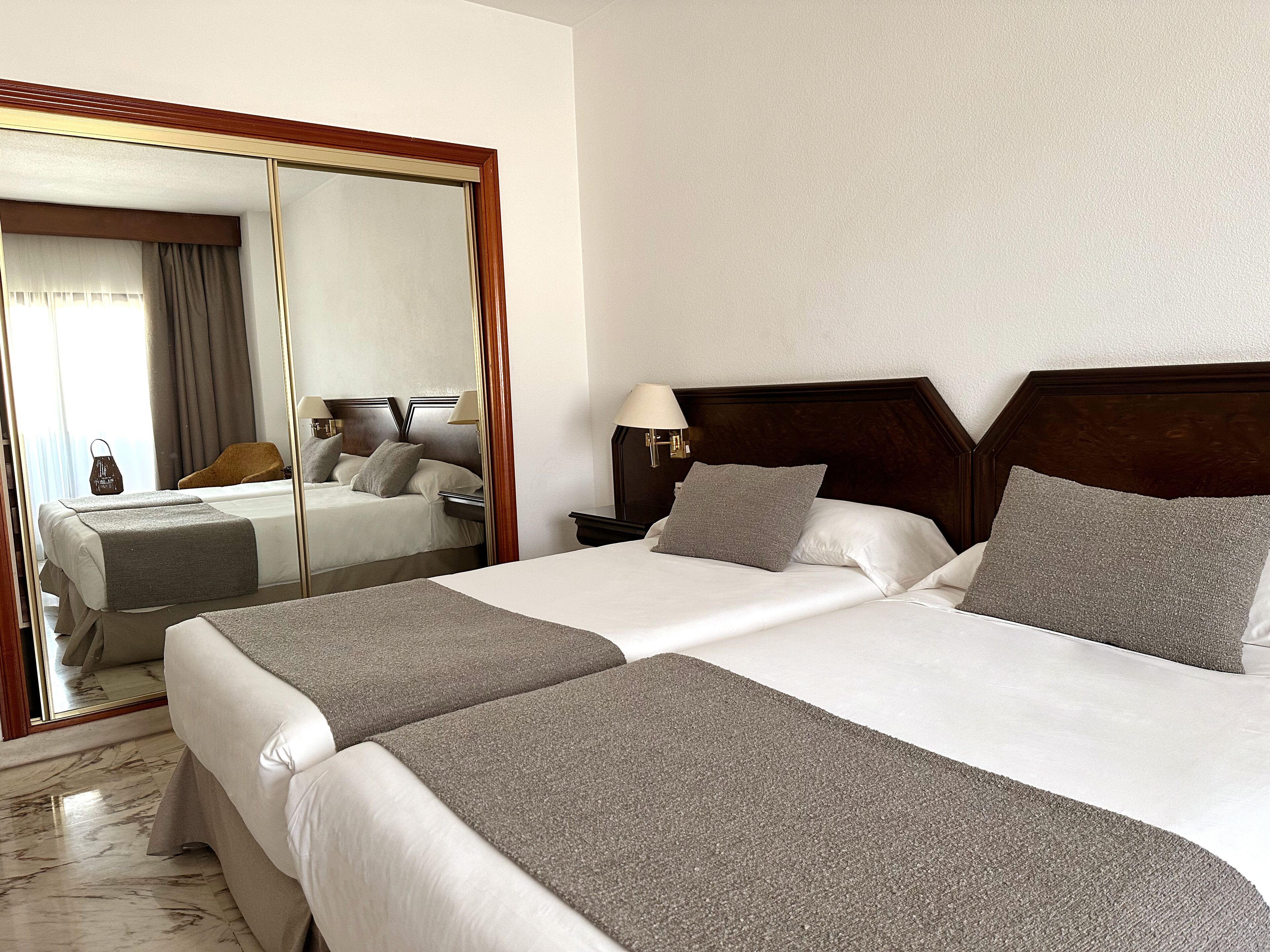 A hotel room with two beds, a large mirror, a nightstand with a lamp, and a view of the city through the window.