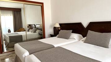 A hotel room with two beds, a large mirror, a nightstand with a lamp, and a view of the city through the window.