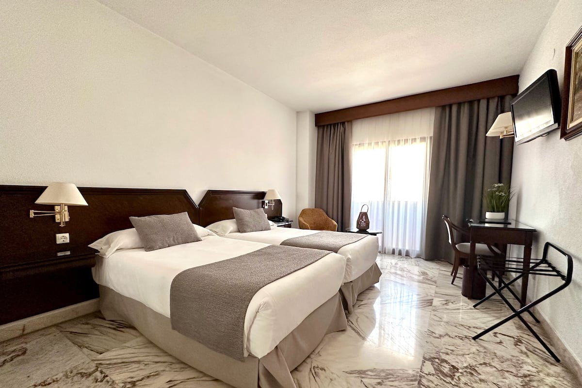 A hotel room with two beds, a desk, a chair, a TV, and a window with curtains.
