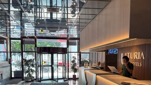 A hotel reception area with a modern design, featuring a reception desk, a digital screen, and a view of the outside through large glass doors. - Hotel Turia Valencia (Valencia)