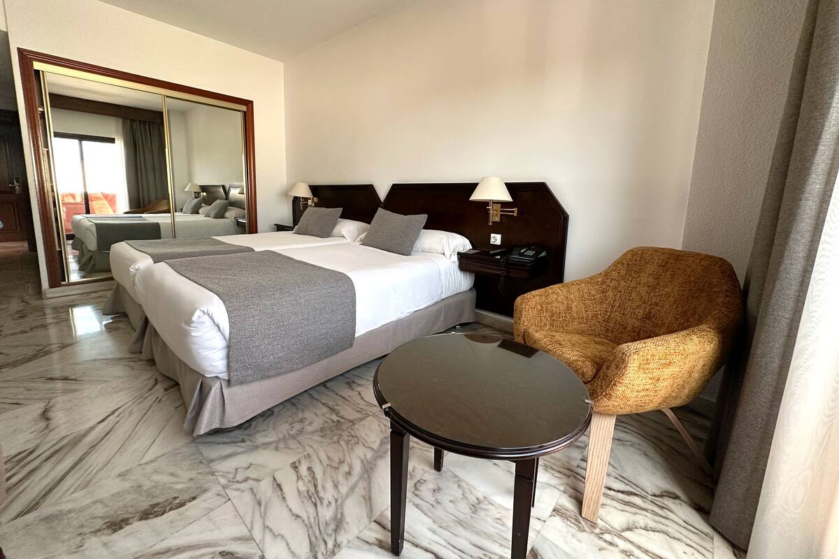A hotel room with two beds, a sitting area with a chair, a small table, and a mirror.