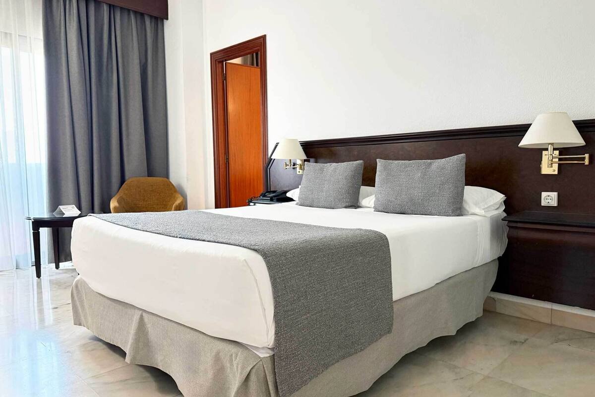A hotel room with a large bed, two bedside lamps, a wooden headboard, a desk with a chair, and a window with curtains.