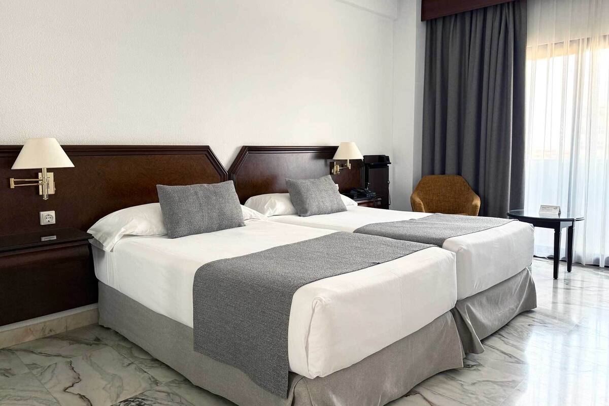 A hotel room with two beds, a nightstand, a chair, a small table, and a window with curtains.