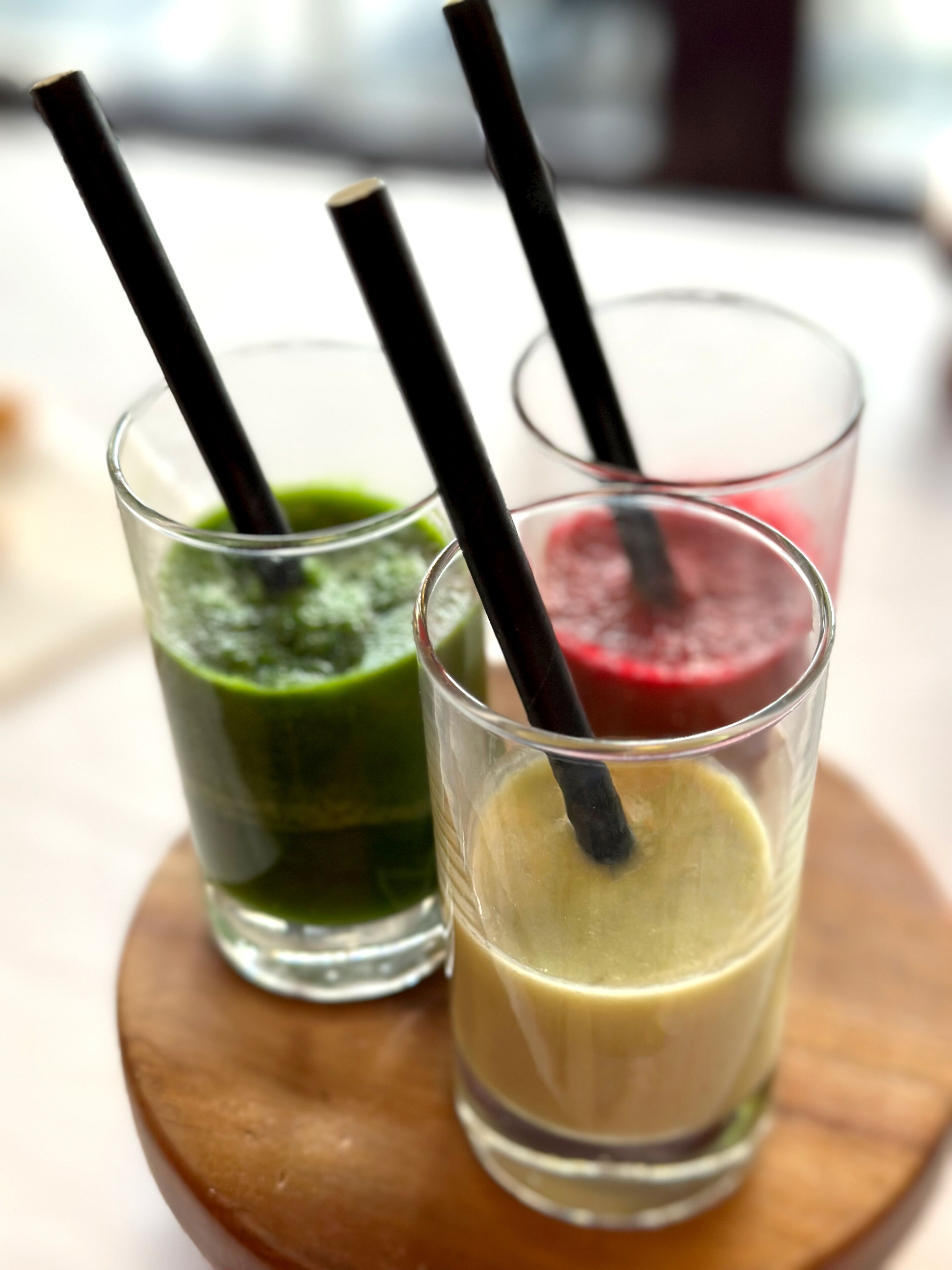 Three glasses of smoothies with black straws on a wooden coaster.