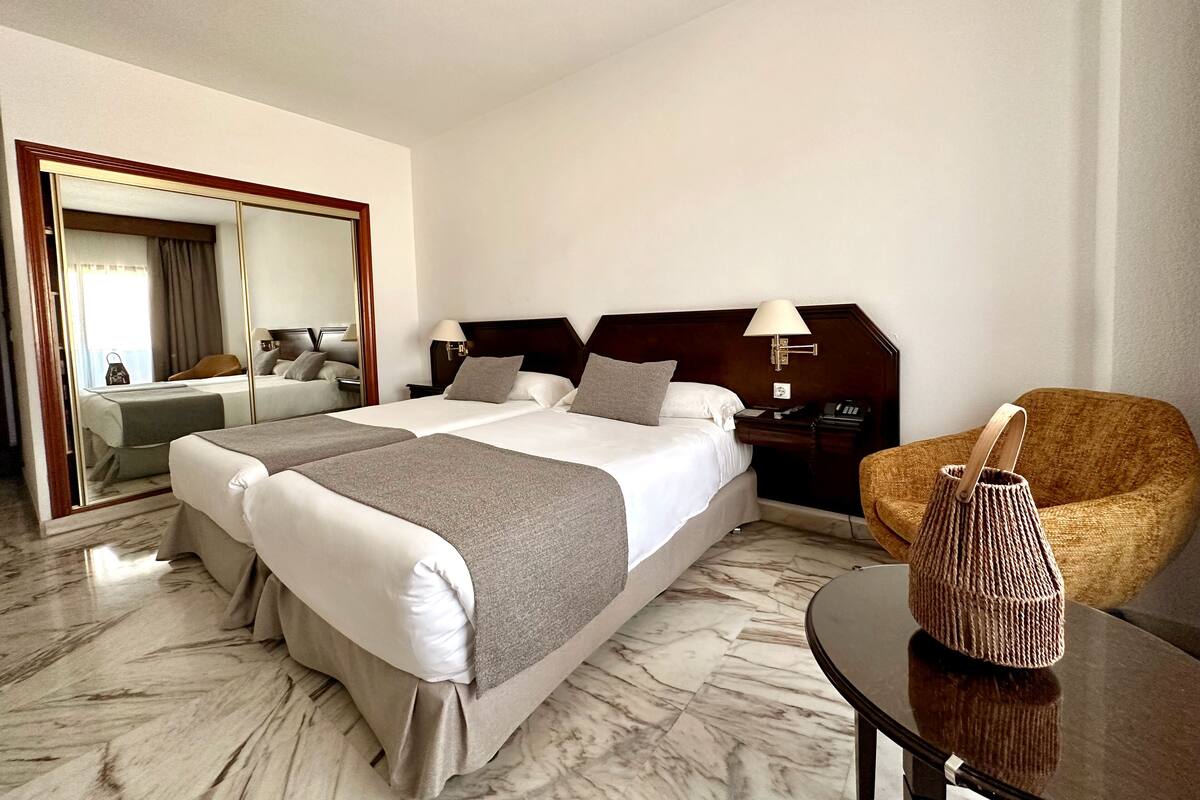 A hotel room with a large bed, a chair, a small table, and a mirror.