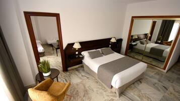 Habitación de hotel con una cama grande, una zona de estar con una silla, una mesita, una planta en maceta y un espejo amplio.