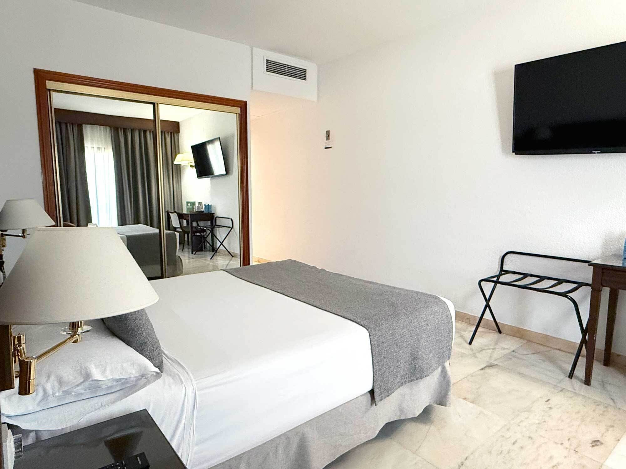 A hotel room with a bed, a desk, a chair, a TV, and a dining area with a table and chairs.