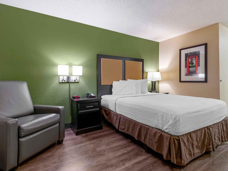 A hotel room with a bed, a chair, a nightstand with a lamp, and a wall-mounted picture.