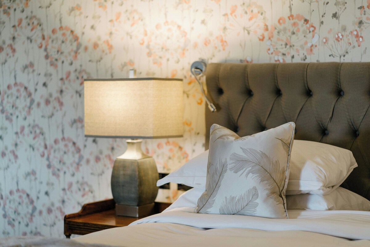 A neatly made bed with a tufted headboard, a bedside lamp, and floral wallpaper.